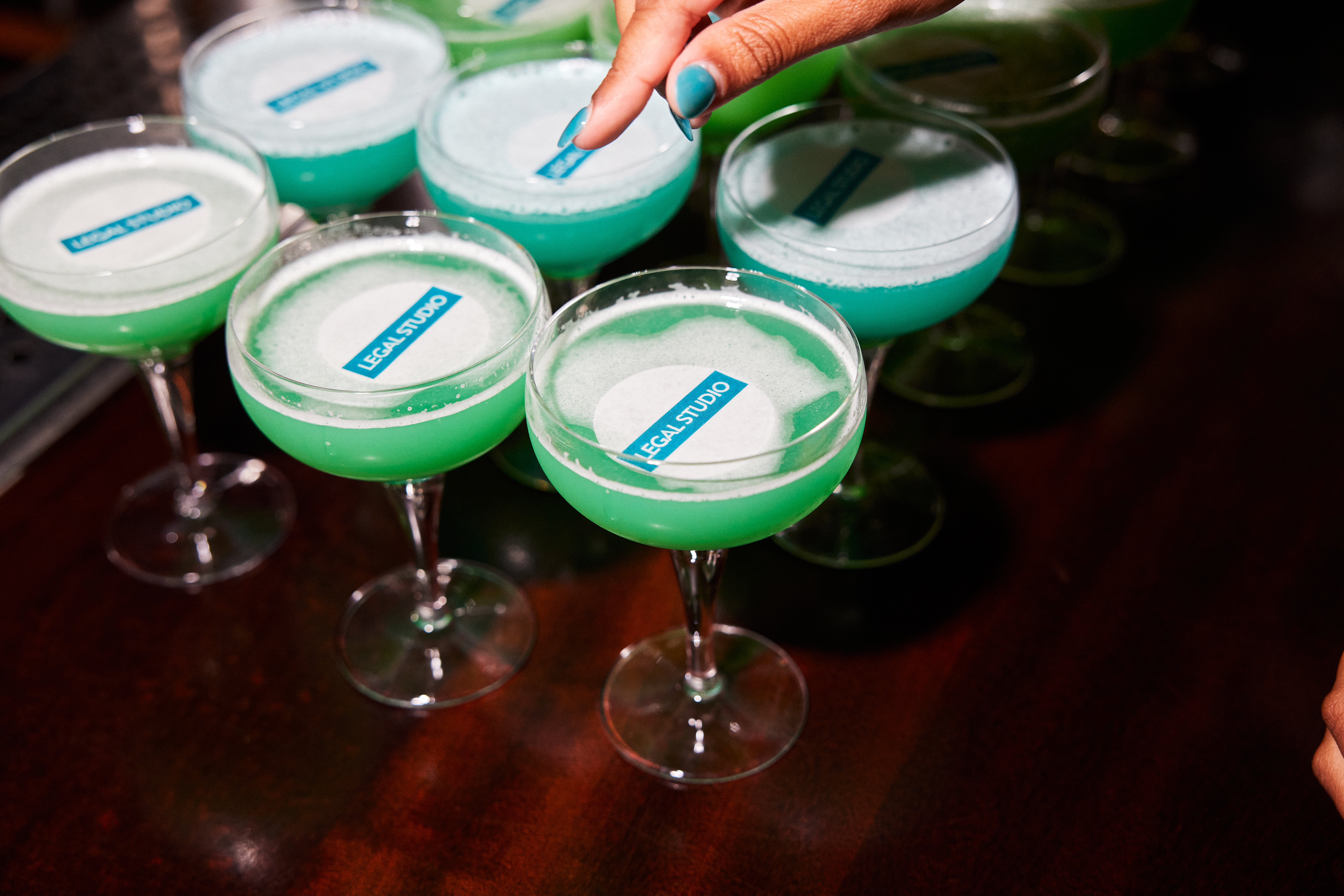 A close-up of several vibrant green cocktails served in coupe glasses, each topped with a circular garnish featuring the Legal Studio logo. A hand with blue-painted nails is seen adding a finishing touch to one of the drinks.