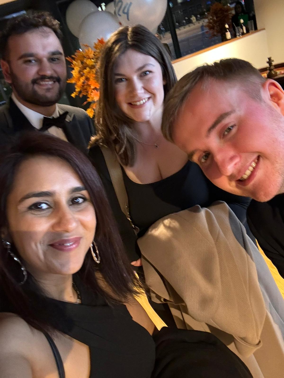 Four people stand together in a group selfie. They are wearing smart evening wear and standing in what appears to be a warmly lit room.